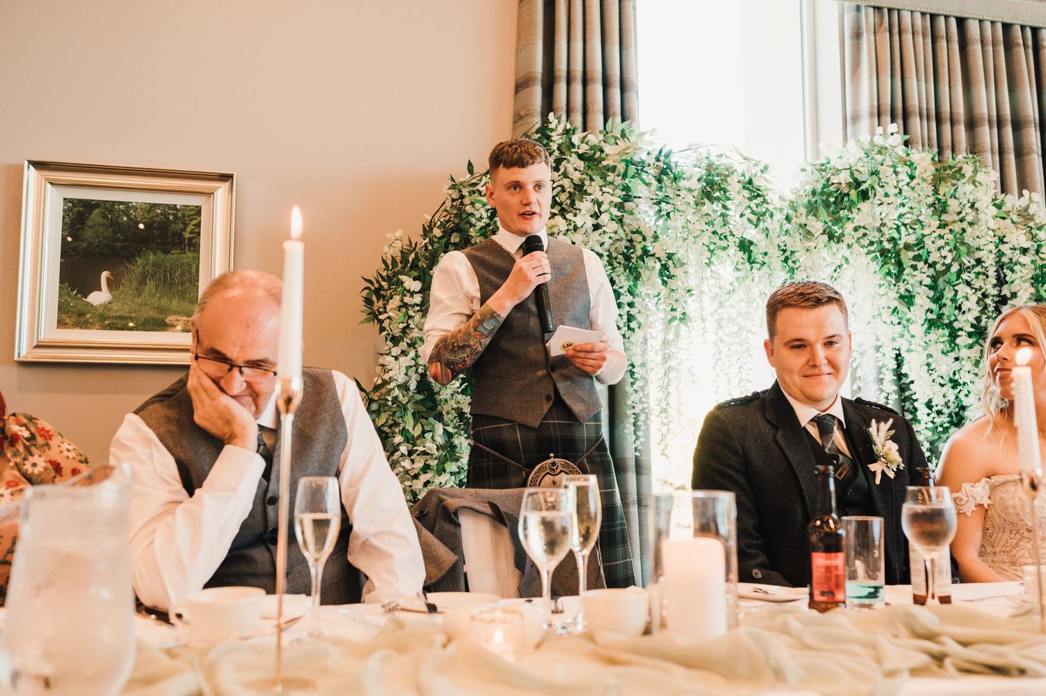 Wedding reception speech moment captured with decorative greenery and candlelit table setting.