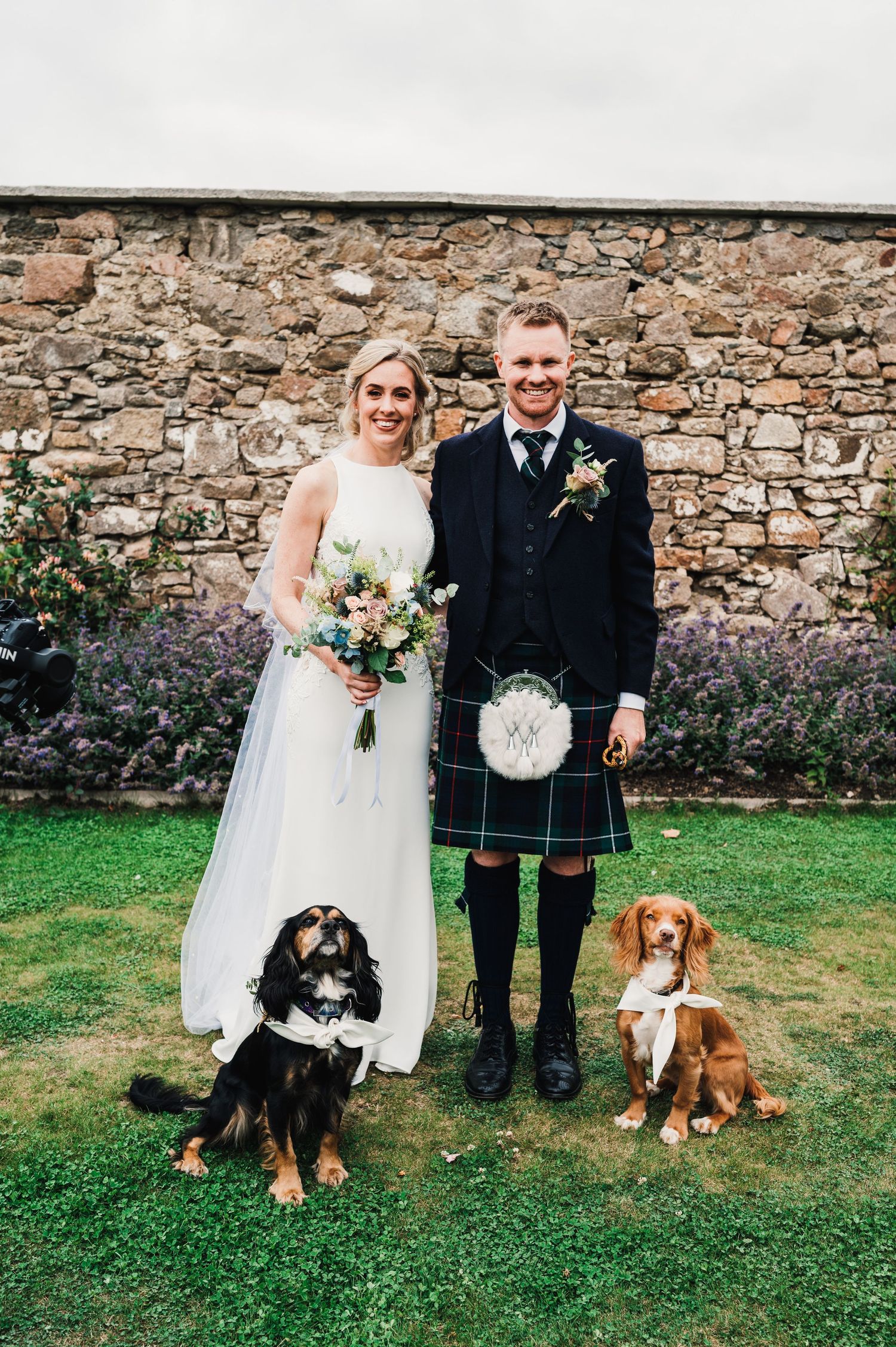 Newlyweds in wedding dress and kilt pose with two dogs against stone wall.