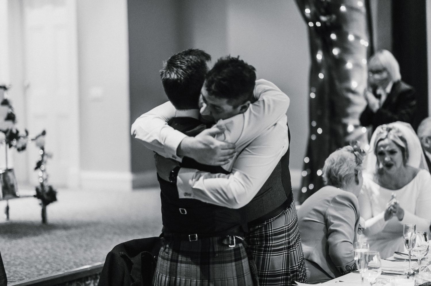 Two wedding guests share an emotional embrace while others watch and laugh during the reception.