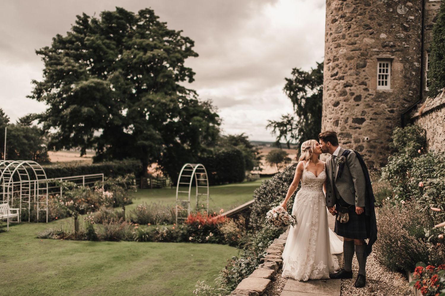 Couple in wedding attire walk along garden path beside castle tower under moody sky.