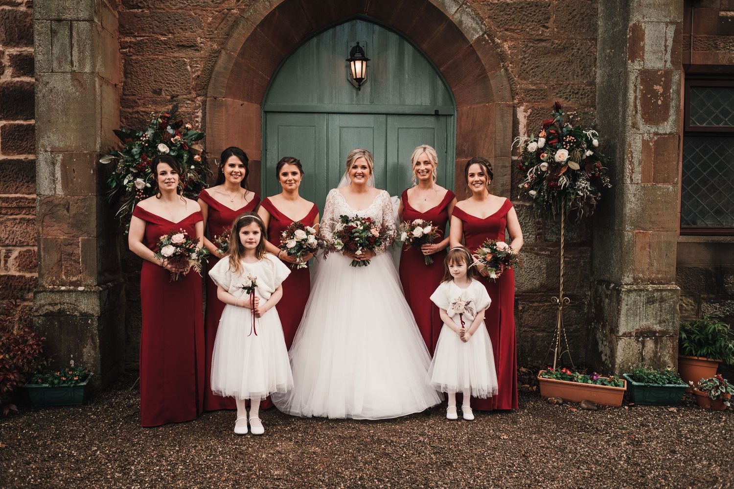 A bride in white dress poses with bridesmaids in burgundy gowns and flower girls at a church entrance.