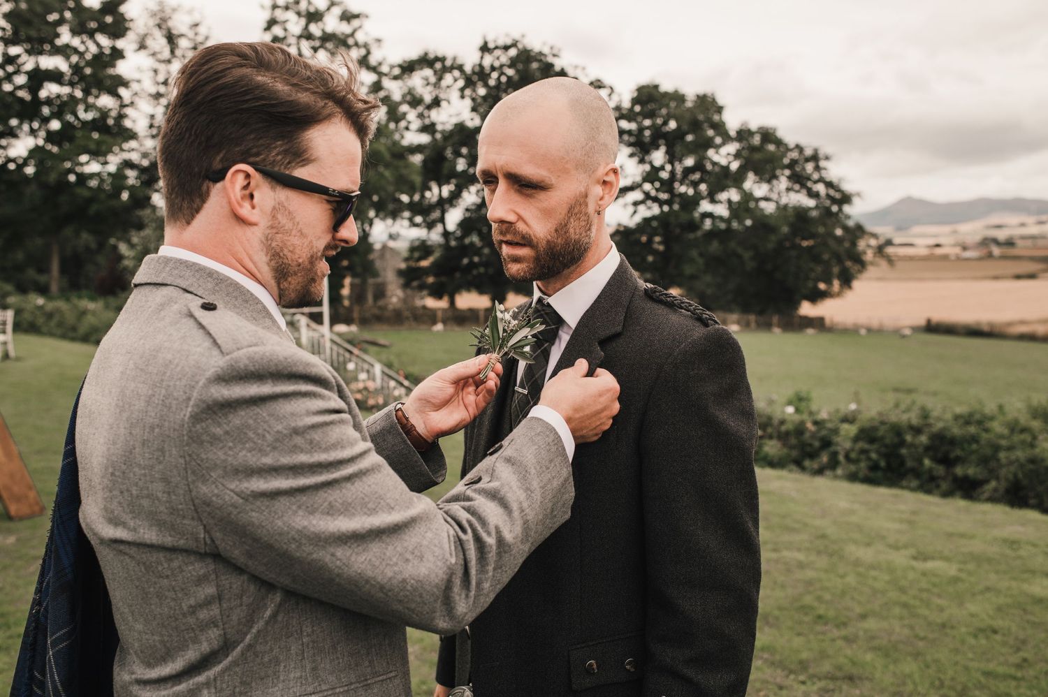 Two men adjusting formal wear and boutonnieres outdoors at wedding event.