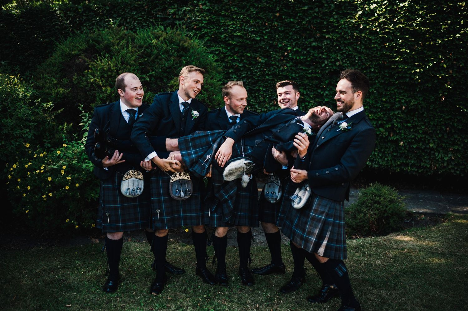 Five groomsmen in traditional Scottish kilts laughing together in garden setting.
