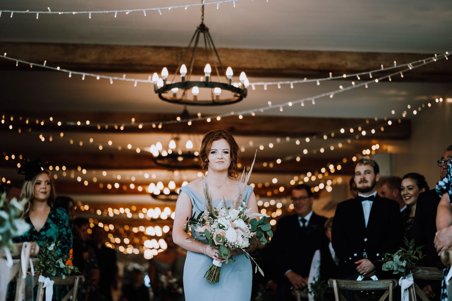 Wedding party member walks down aisle under romantic string lights and chandeliers.