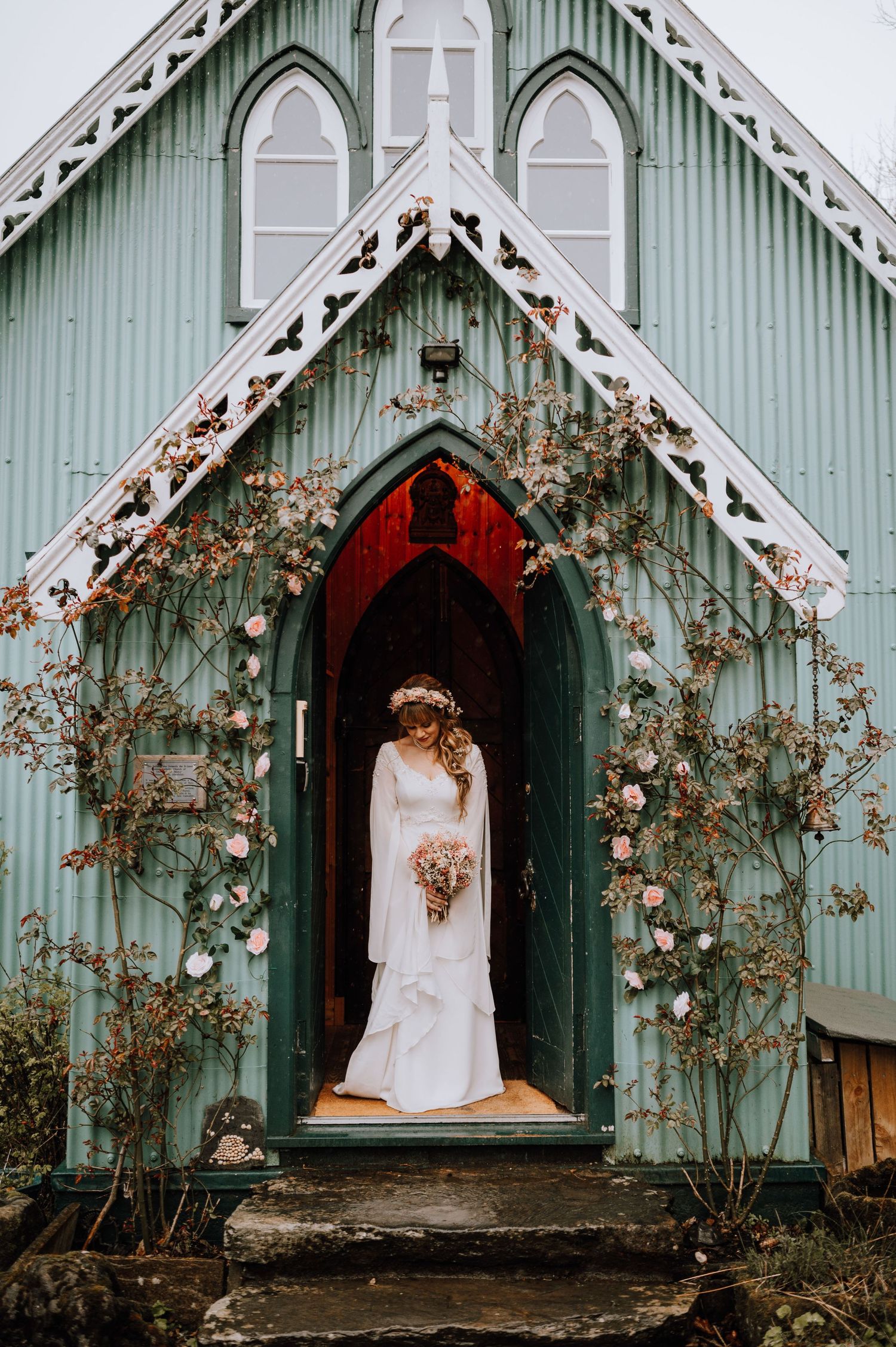 Bride in white gown stands in doorway of mint green chapel decorated with climbing roses.