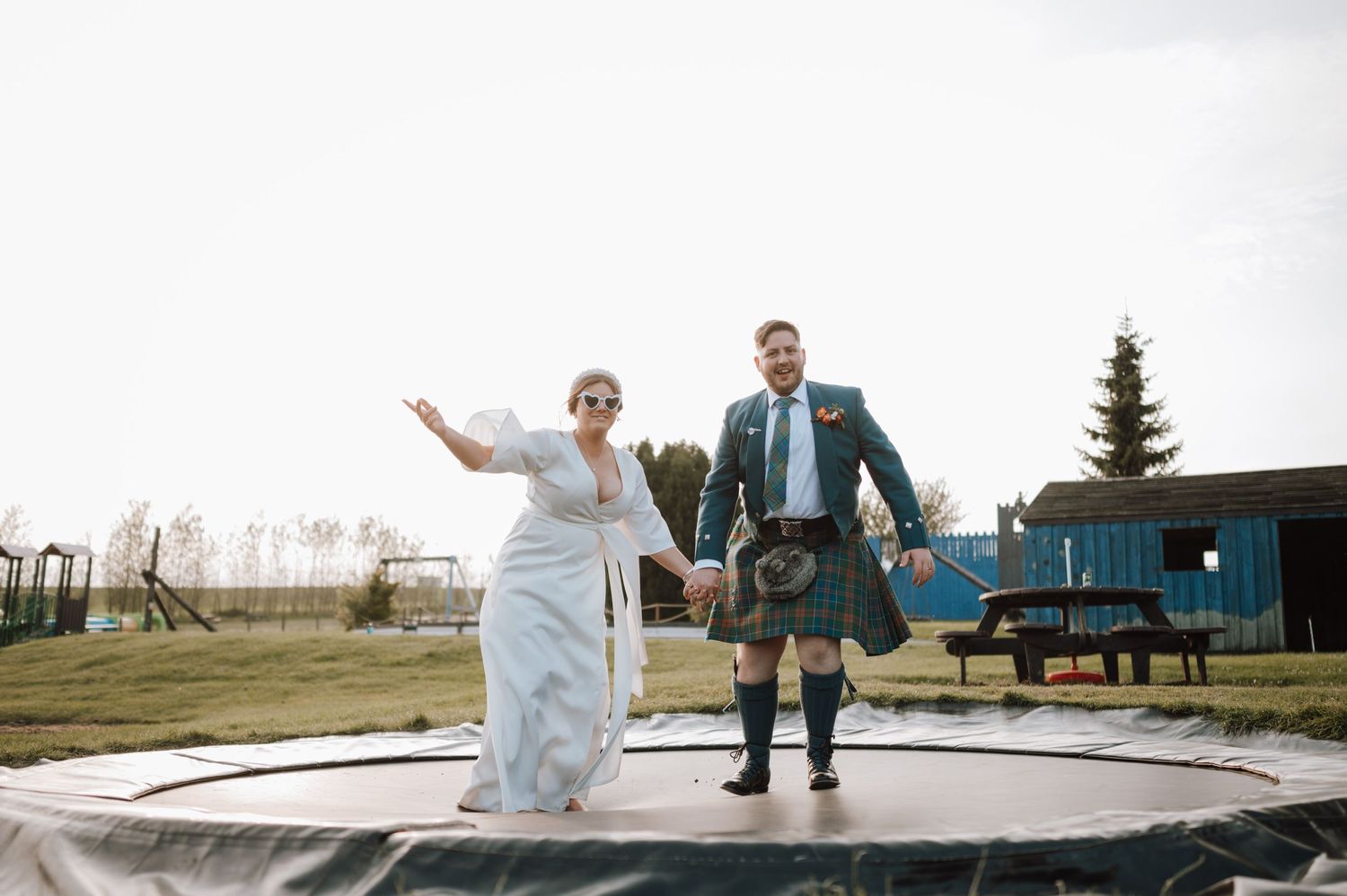 Wedding couple jumps playfully on a trampoline at an outdoor venue with the groom wearing a traditional kilt.