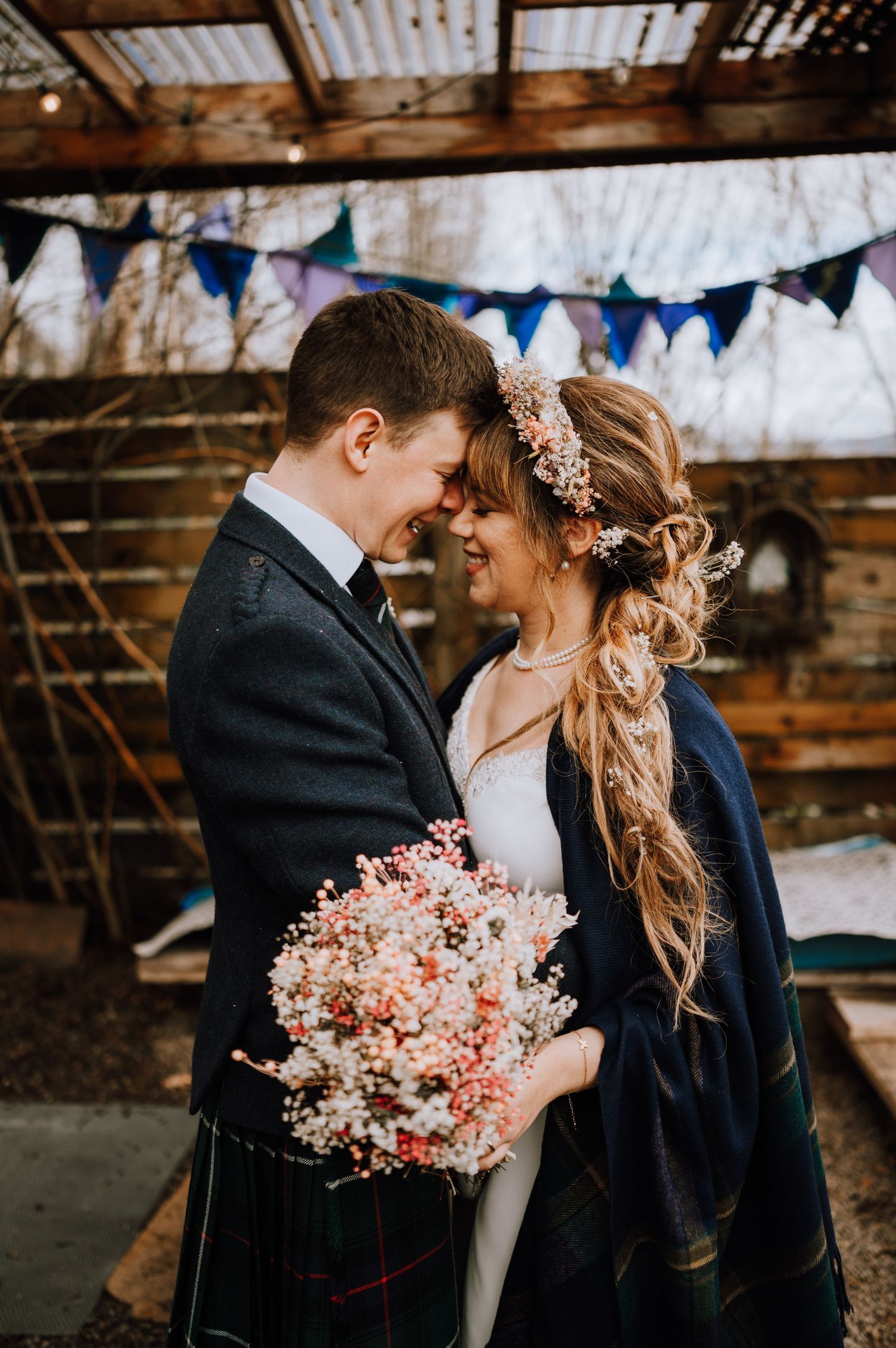 Romantic wedding scene with blue bunting, pink flowers, and Scottish wedding attire in a rustic setting.