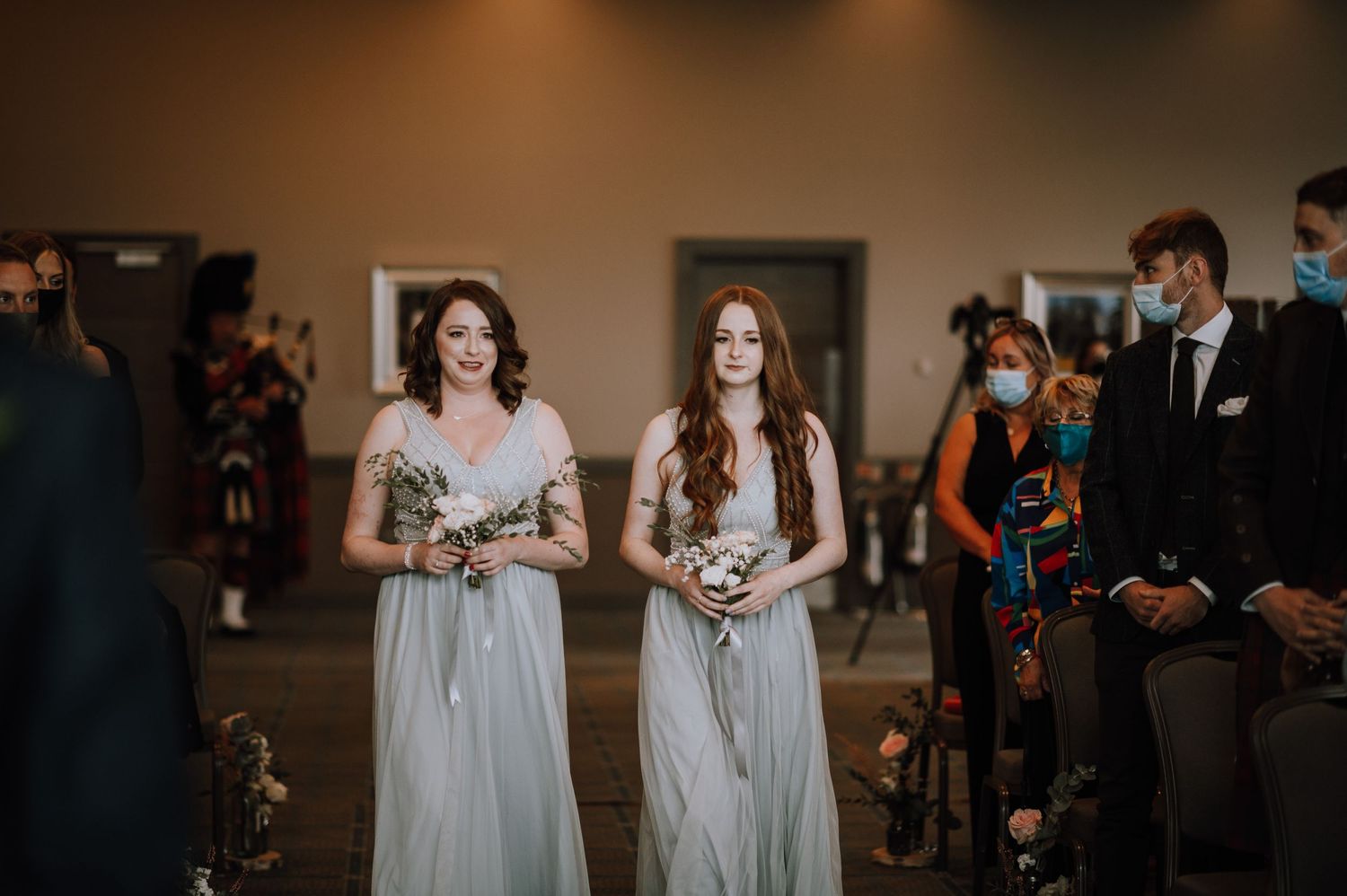 Bridesmaids in pale grey dresses walk down aisle during indoor ceremony with seated guests.