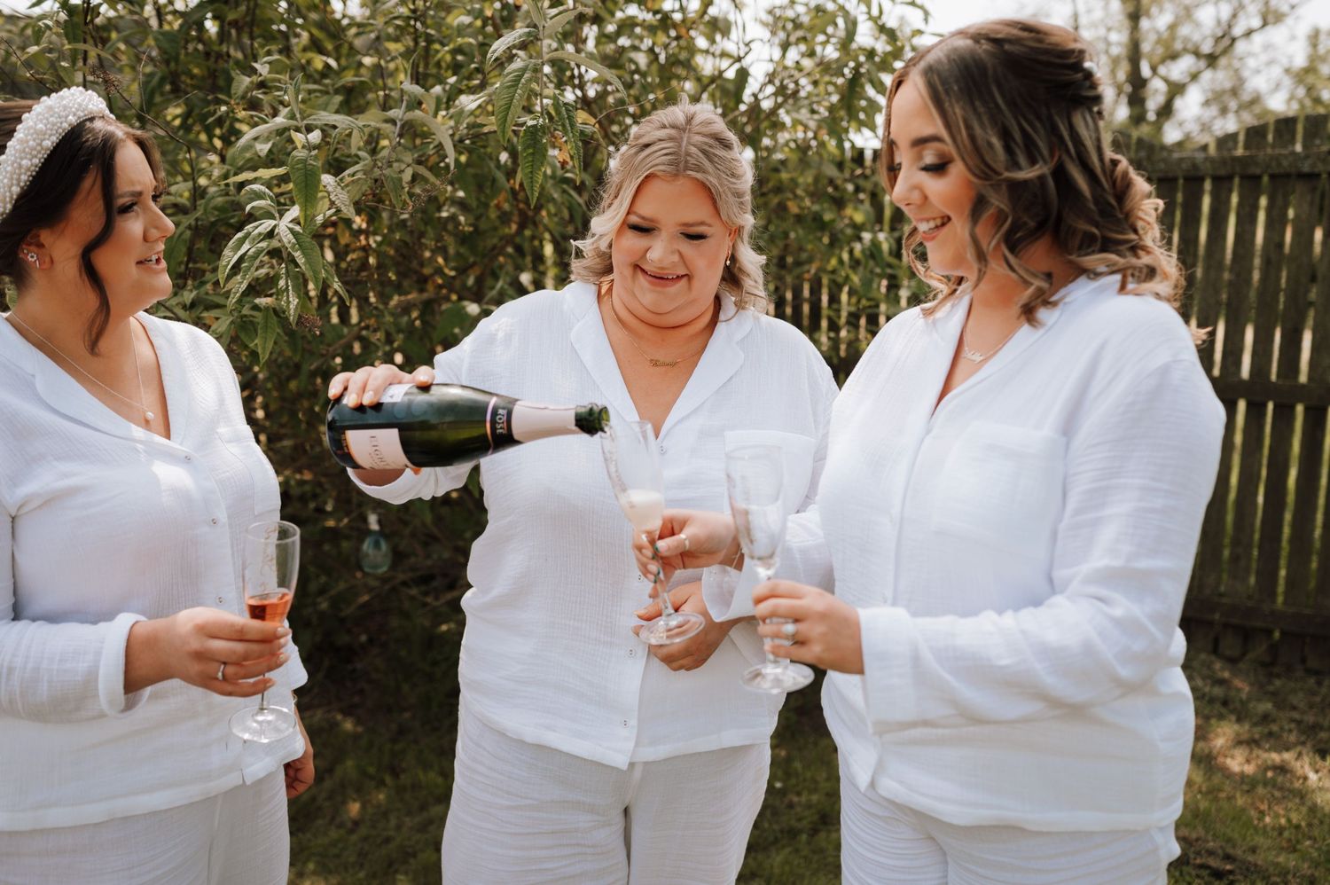Three people in white outfits sharing champagne and laughter in an outdoor garden setting.