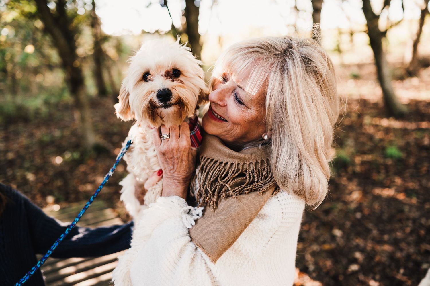 A small white dog shares affectionate moments with someone during a golden hour outdoor photoshoot in a wooded setting.