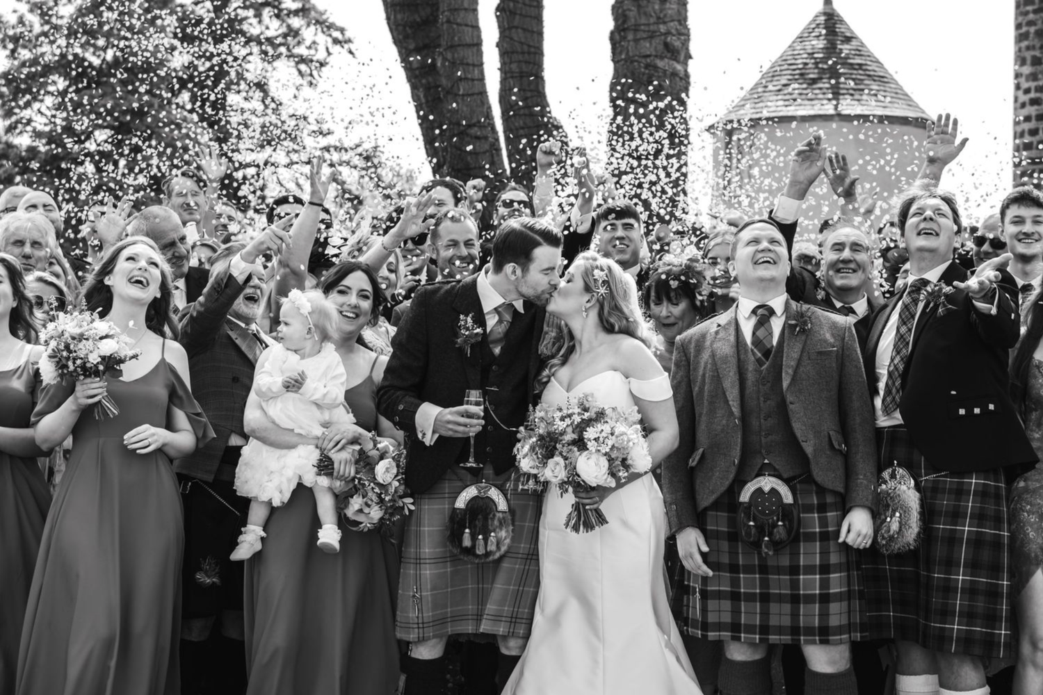 A joyful wedding party stands in a line outdoors with trees in the background during a celebration.