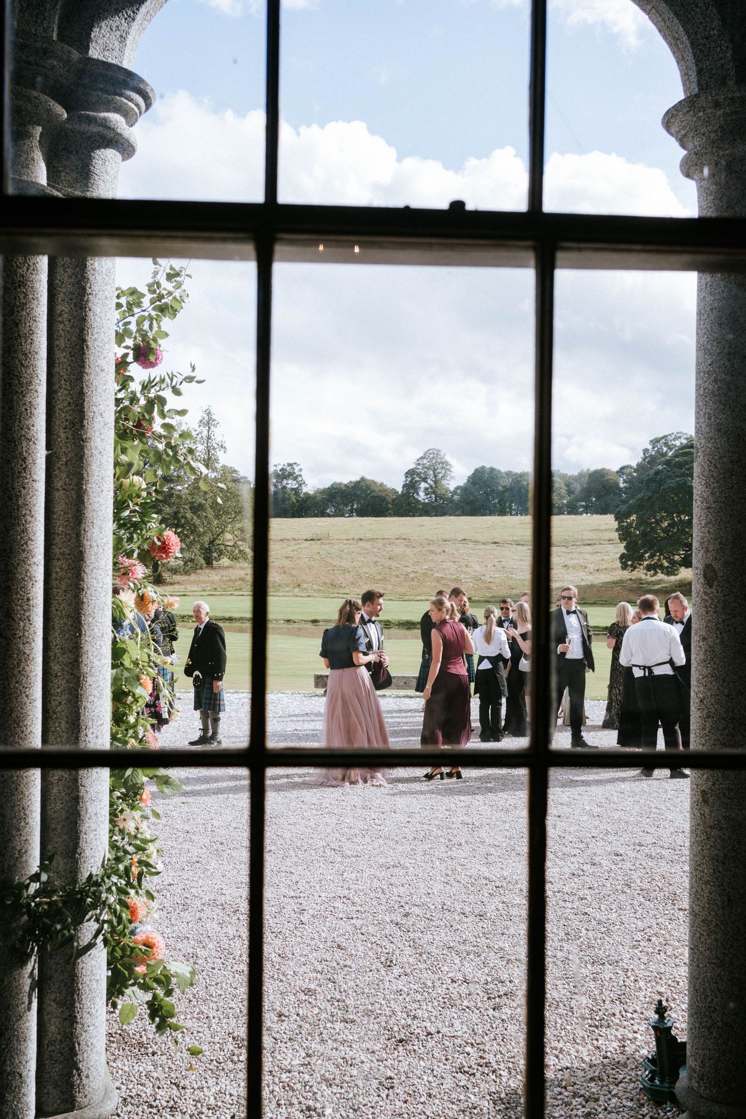 View through an arched window showing guests gathering on a gravel path with rolling grounds beyond.