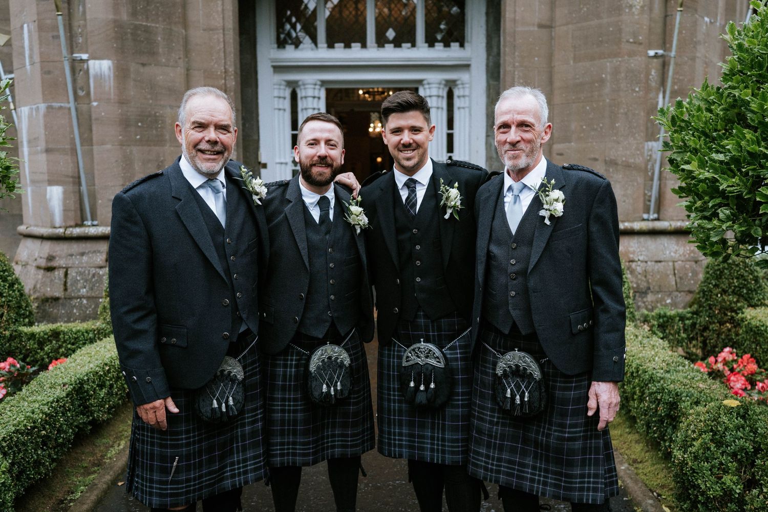 Wedding party wearing traditional Scottish kilts and formal attire posing outside a stone church.