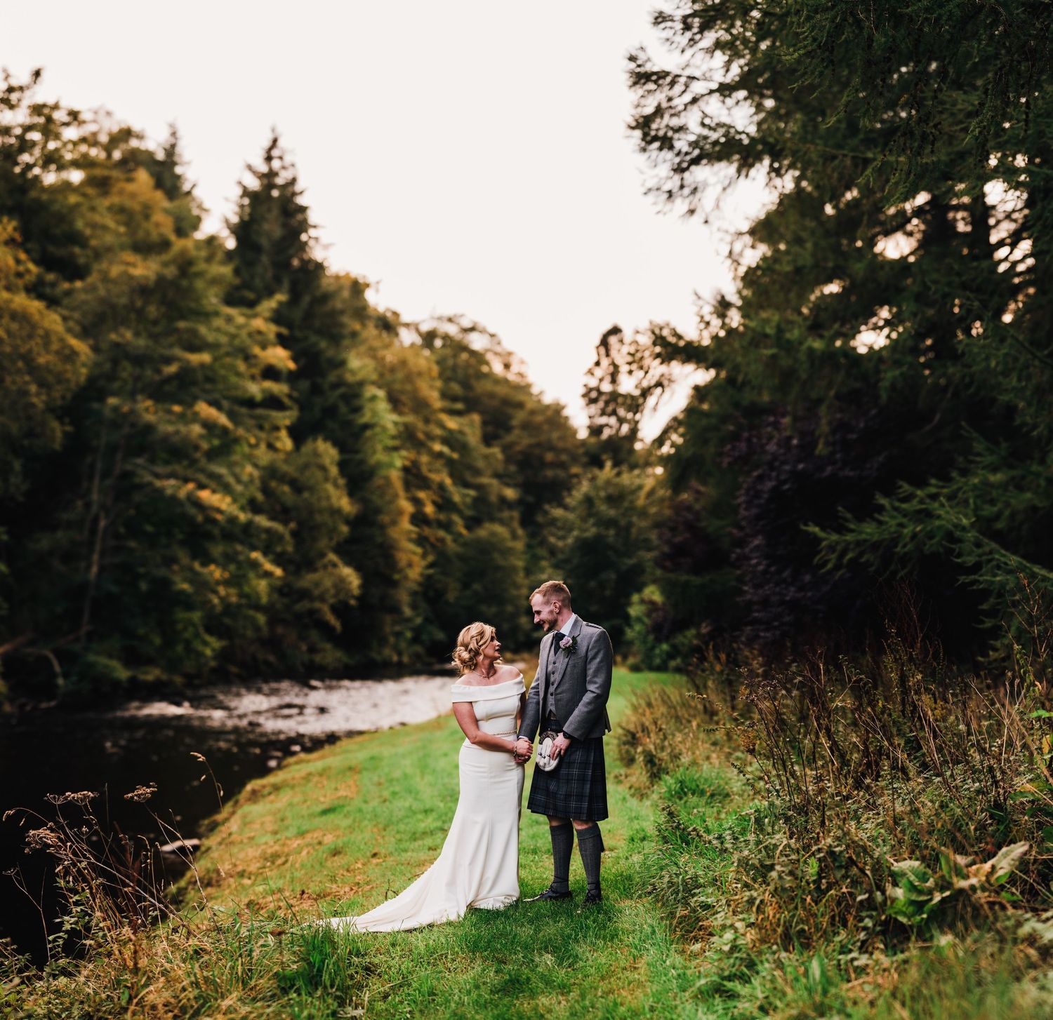 A romantic couple walks along a scenic path surrounded by lush greenery and trees during golden hour.