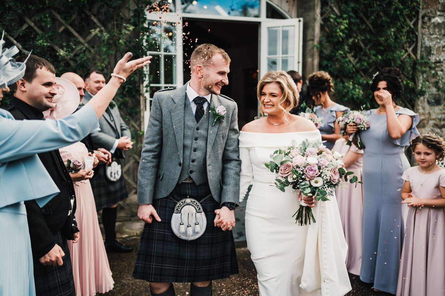 Wedding party celebrates outdoors with pink bridesmaid dresses and traditional Scottish attire.