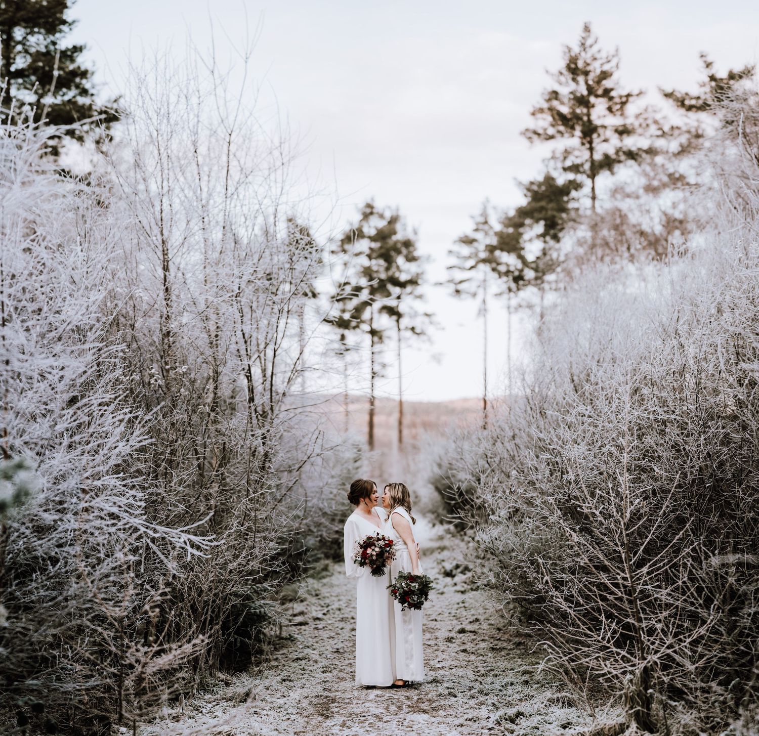 Same Sex Wedding Photography. Two people in white dresses stand together on a snowy winter path surrounded by frost-covered trees.