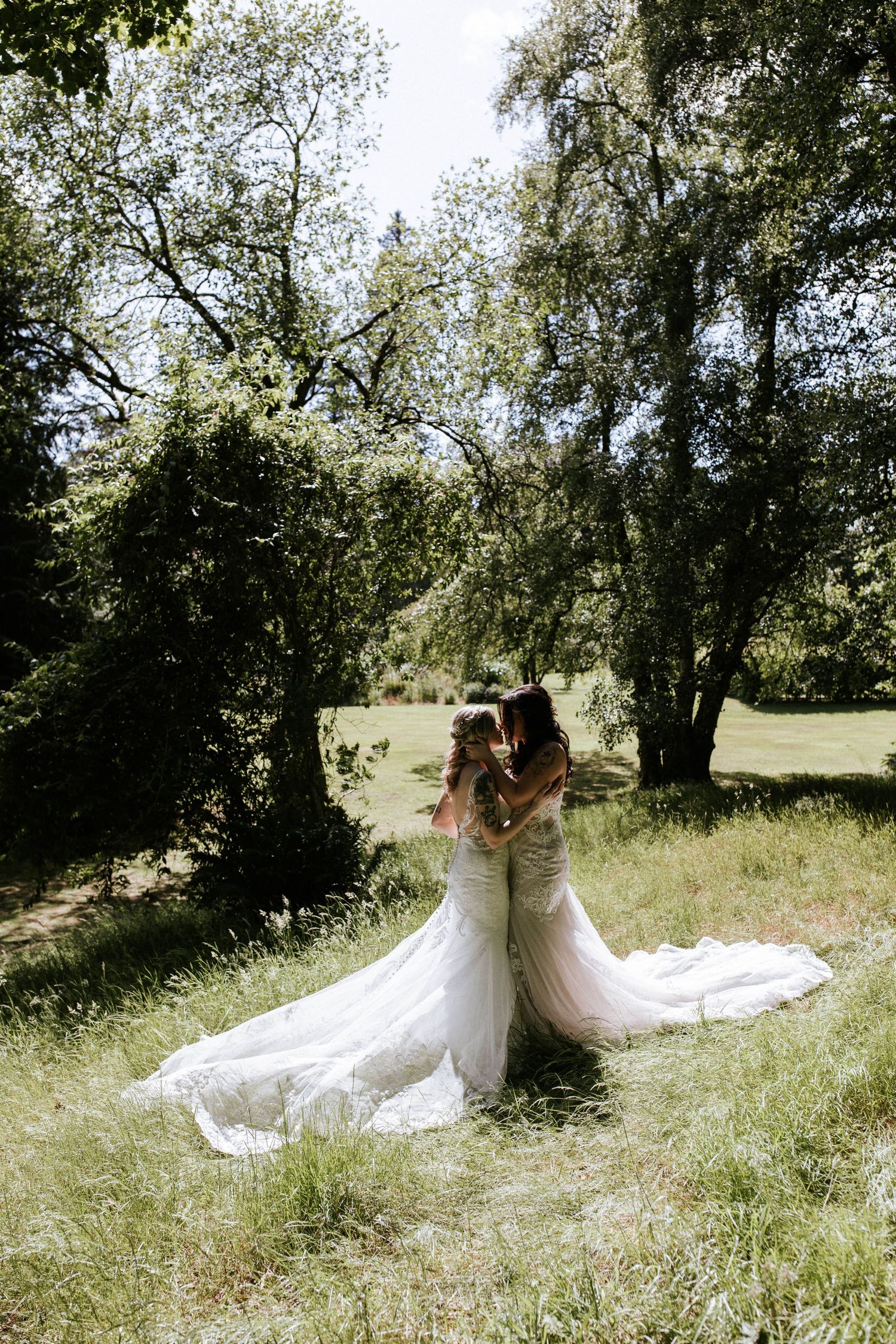 Two people in flowing white wedding gowns pose together in a sunny meadow with trees.