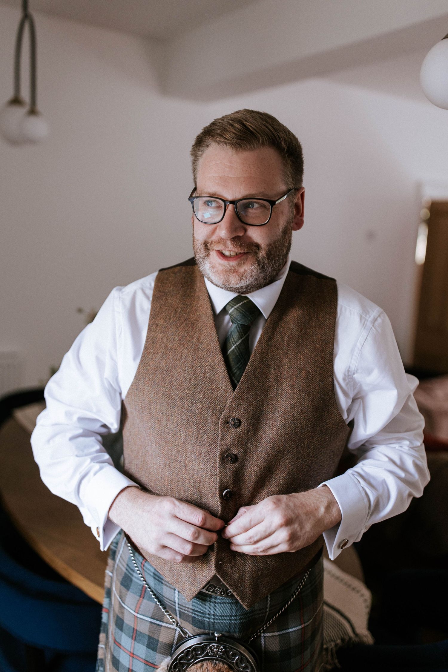 Person wearing a brown tweed vest and white shirt buttoning up formal attire.
