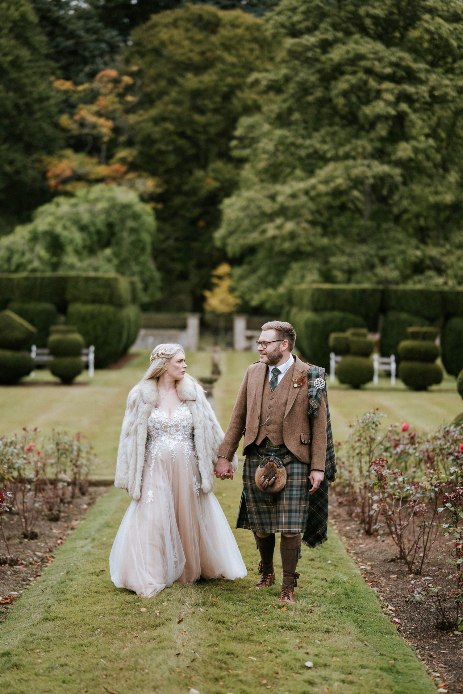 A couple walks through a garden path lined with rose bushes on their wedding day.