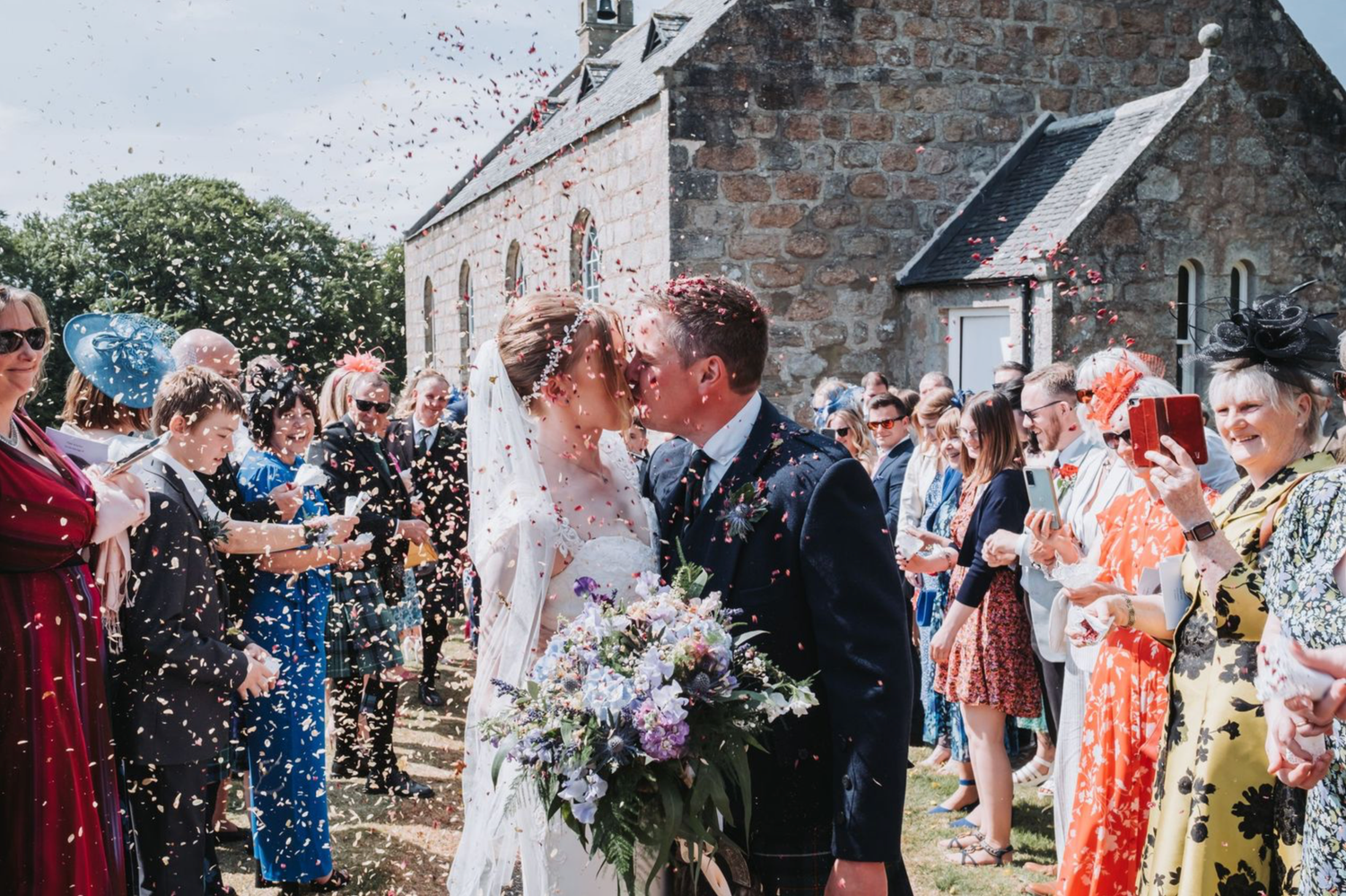 Wedding guests shower newlyweds with confetti outside a stone church on a sunny summer day.