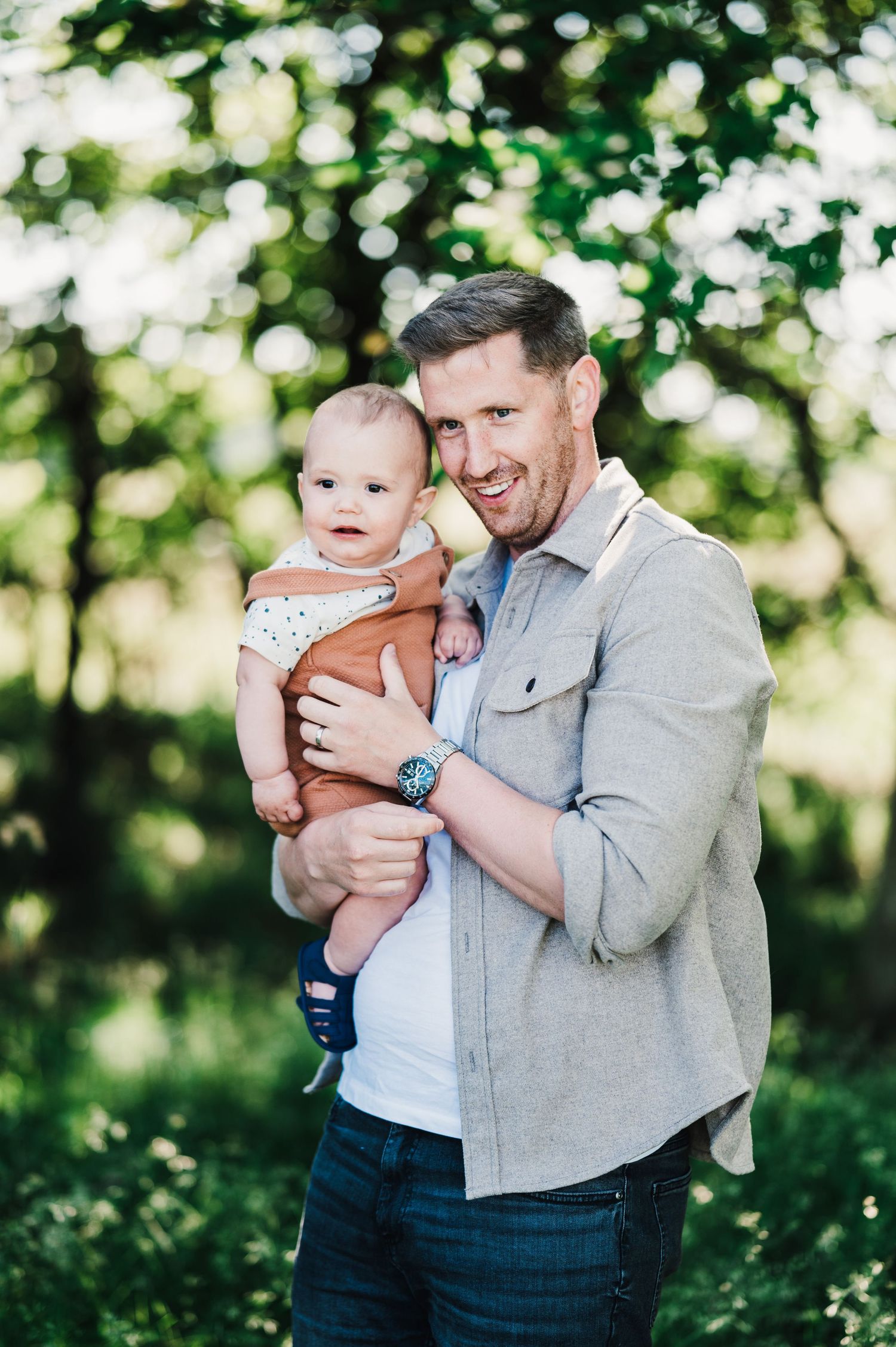 A baby in a striped onesie is held outdoors by someone wearing a gray jacket against a blurred green nature backdrop.
