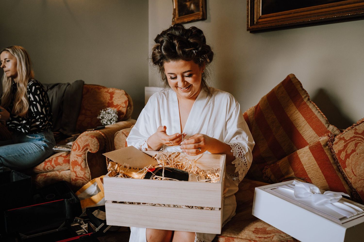 A person in a white robe opens a gift box while sitting on a brown couch during wedding preparations.