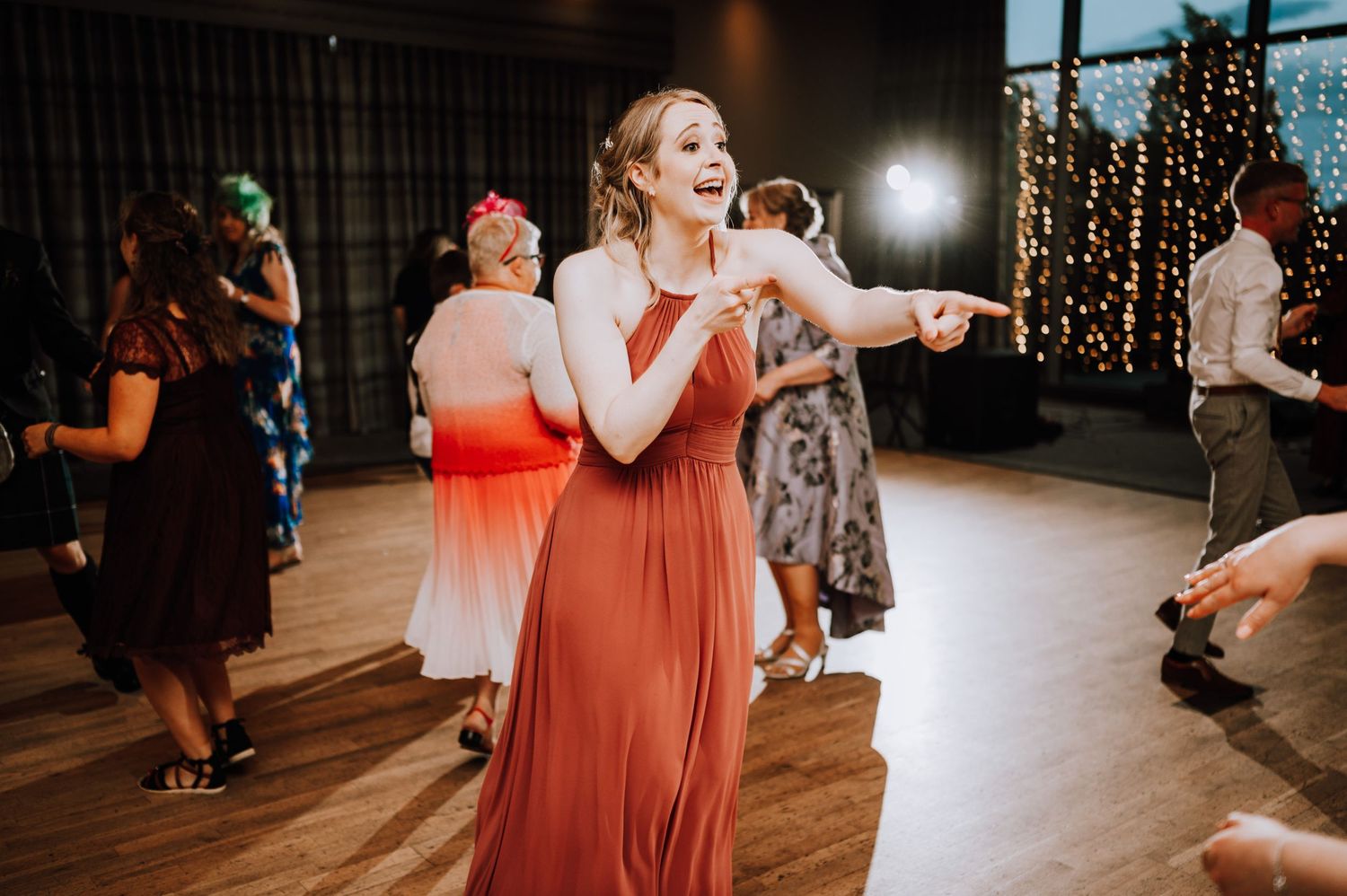 A person in a coral-colored dress dances enthusiastically on a wooden dance floor under string lights.