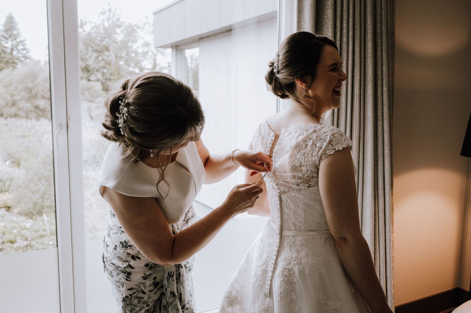 Wedding preparations showing final dress adjustments near a bright window with elegant lace detailing.