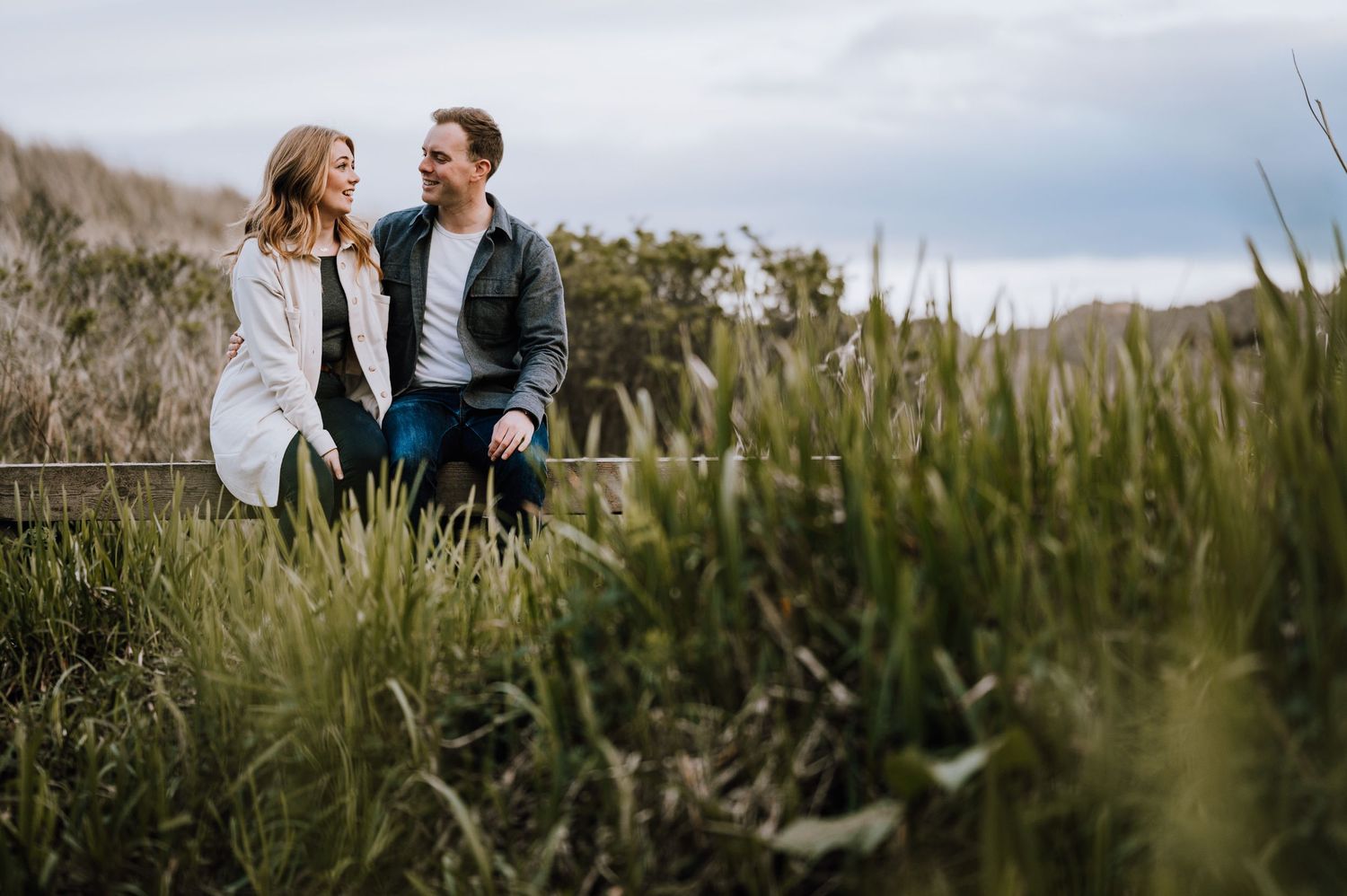 A couple sits intimately together on a wooden bench amid tall coastal grasses.
