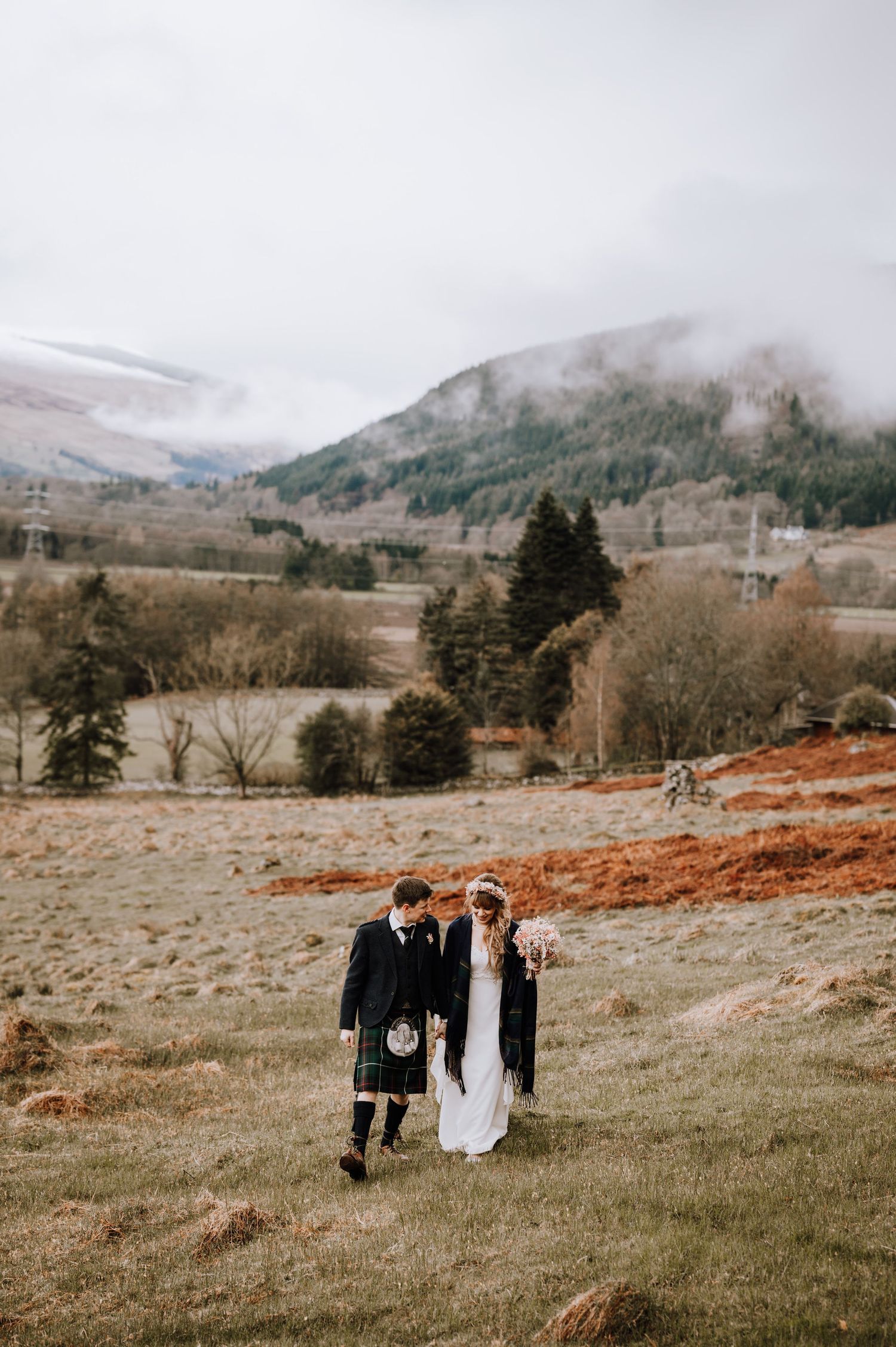 Couple in wedding attire walks through foggy Scottish Highland meadow with mountains beyond.