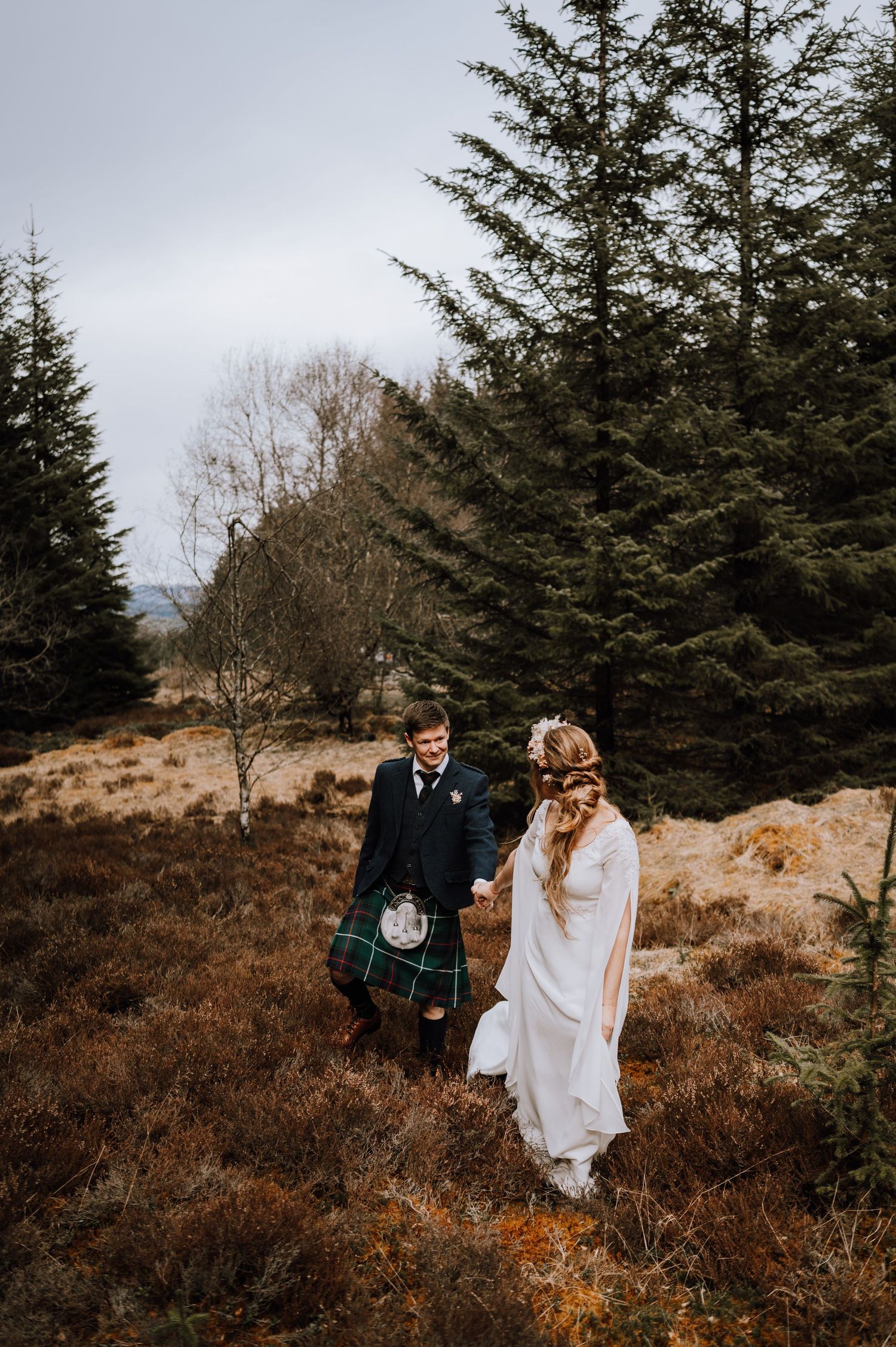 Traditional Scottish wedding couple standing among pine trees on a misty Highland hillside.