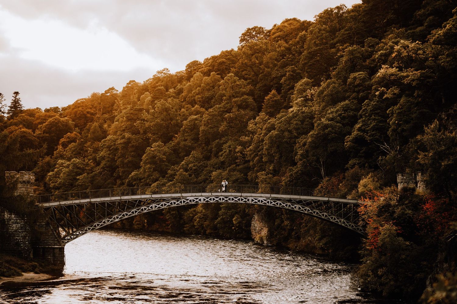 Scenic arched bridge crossing river surrounded by dense autumn forest at sunset.