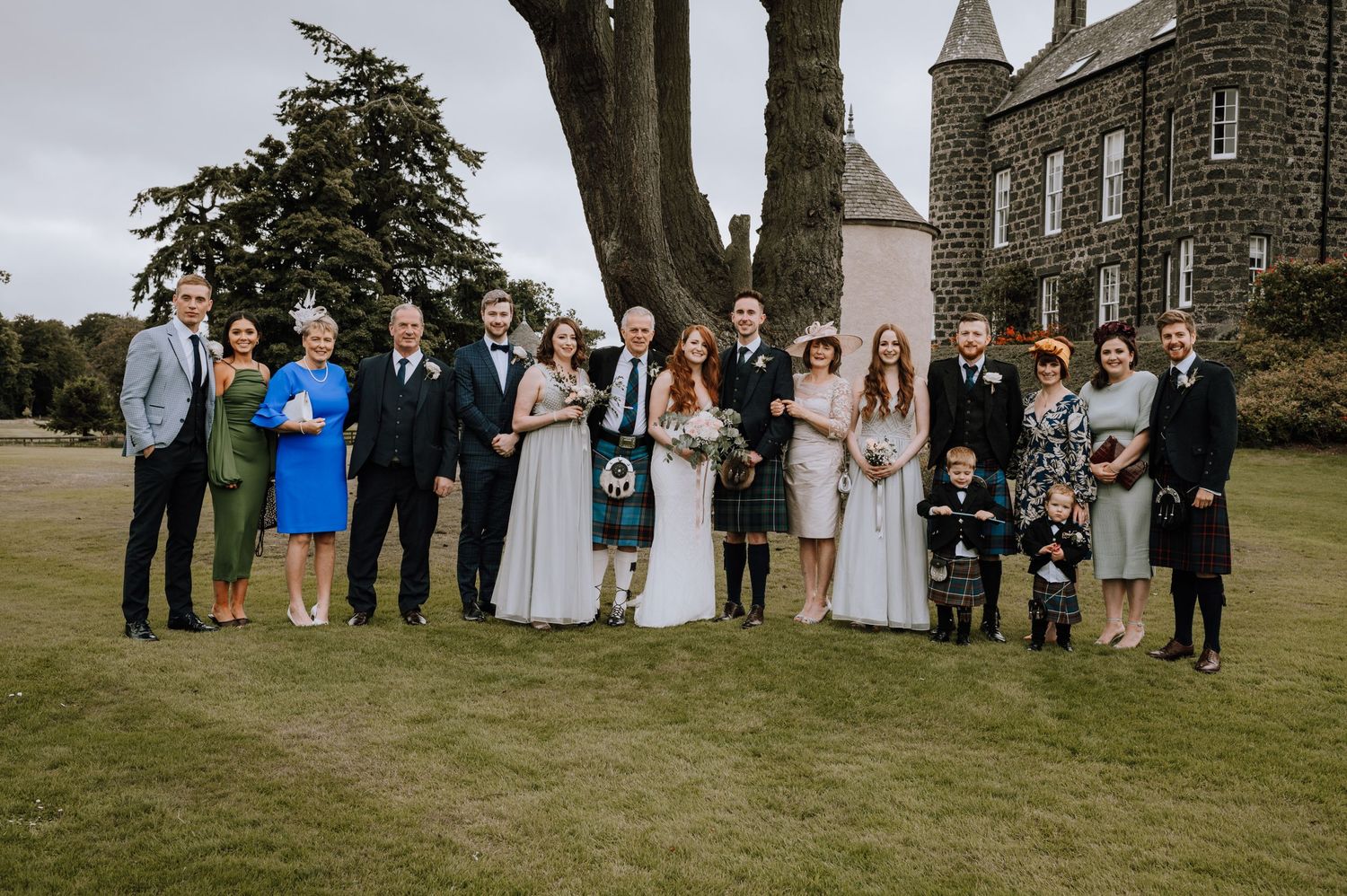 Wedding party gathered on lawn in front of stone Scottish castle with traditional kilts and formal attire.