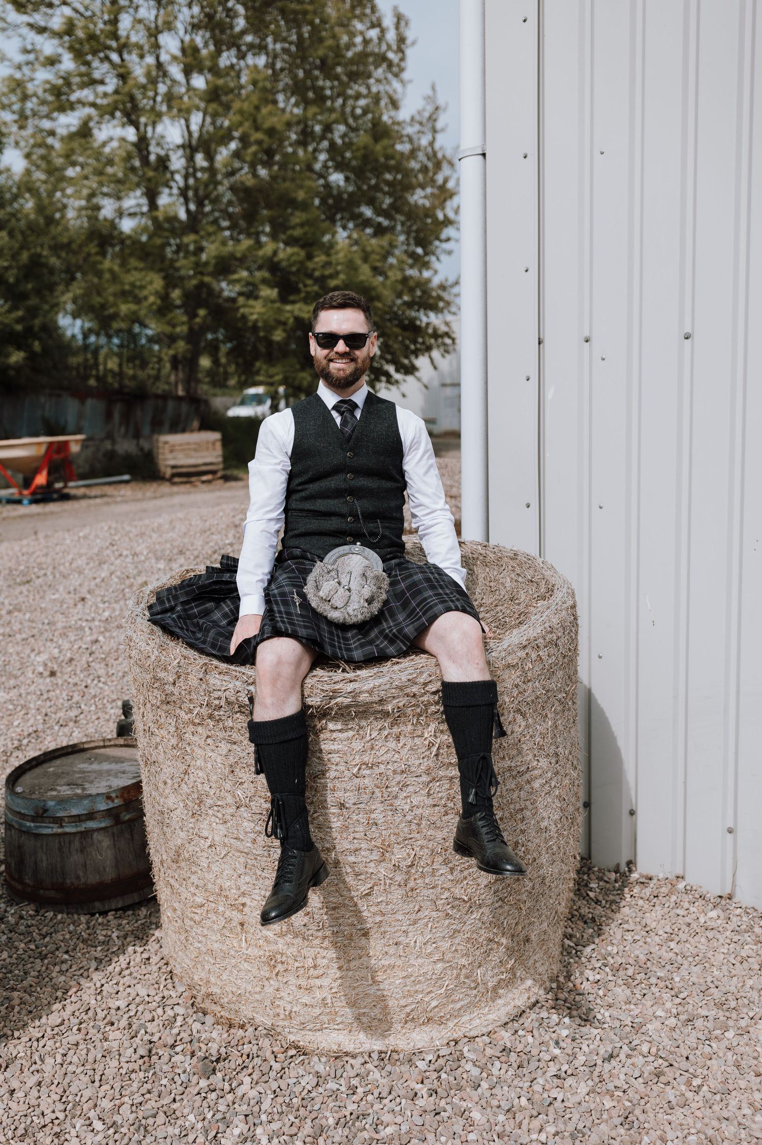 Person wearing traditional Scottish kilt and vest sits on concrete structure outdoors.