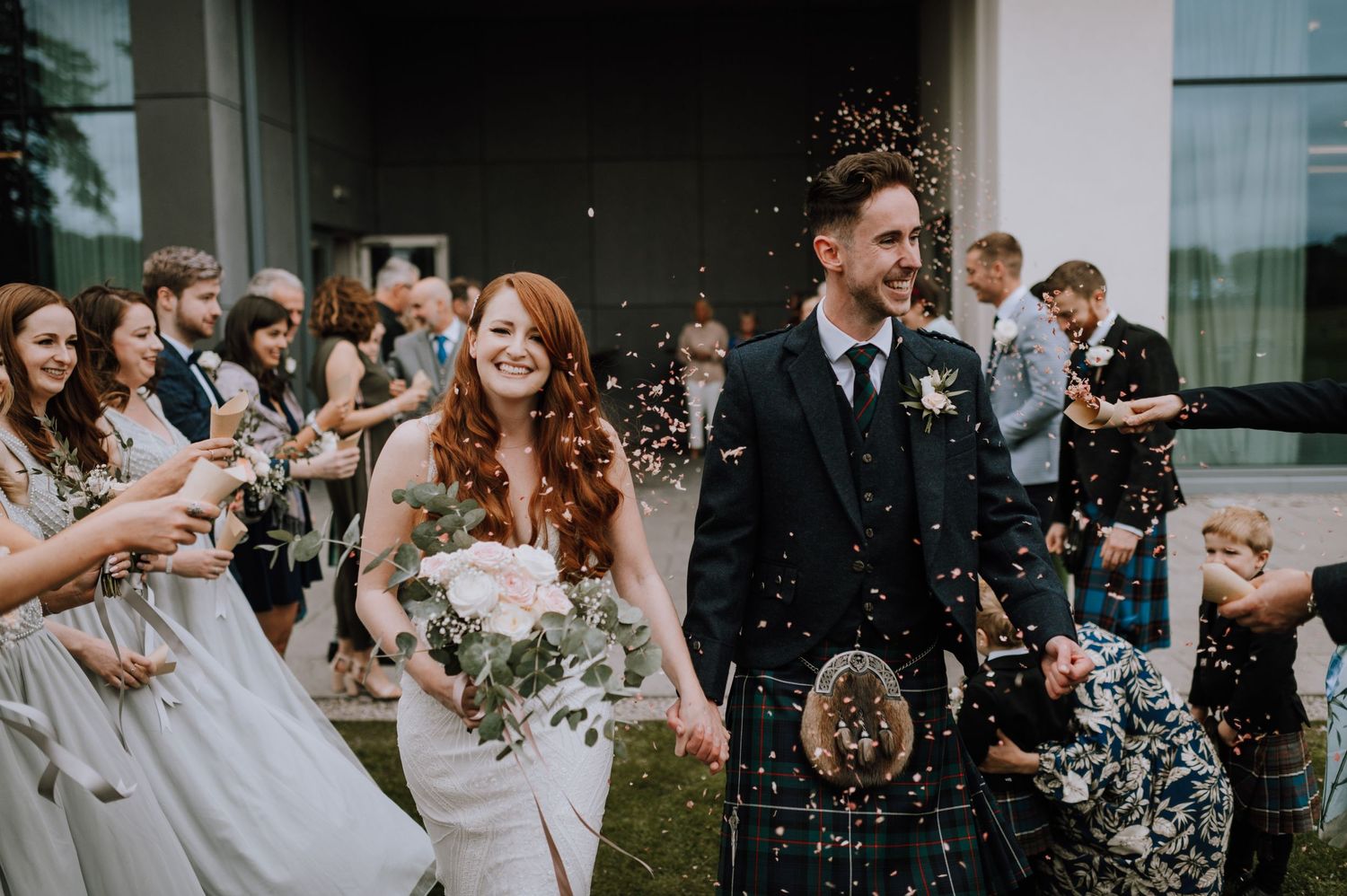 Guests throw confetti at a Scottish wedding ceremony with the groom wearing a traditional kilt.