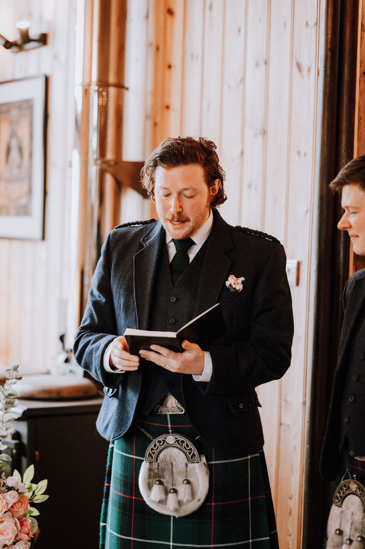 A person in formal attire and traditional Scottish kilt reads from a book during a ceremony.