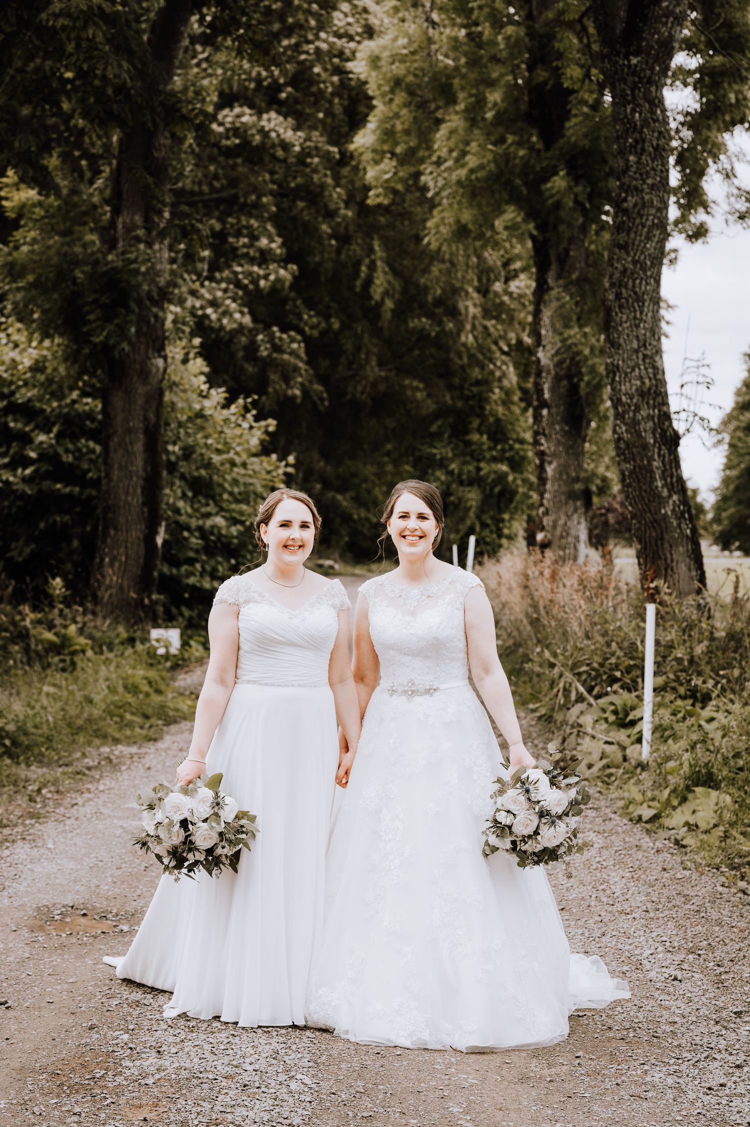 Two brides in white wedding dresses hold beautiful bouquets while standing on a tree-lined gravel path.