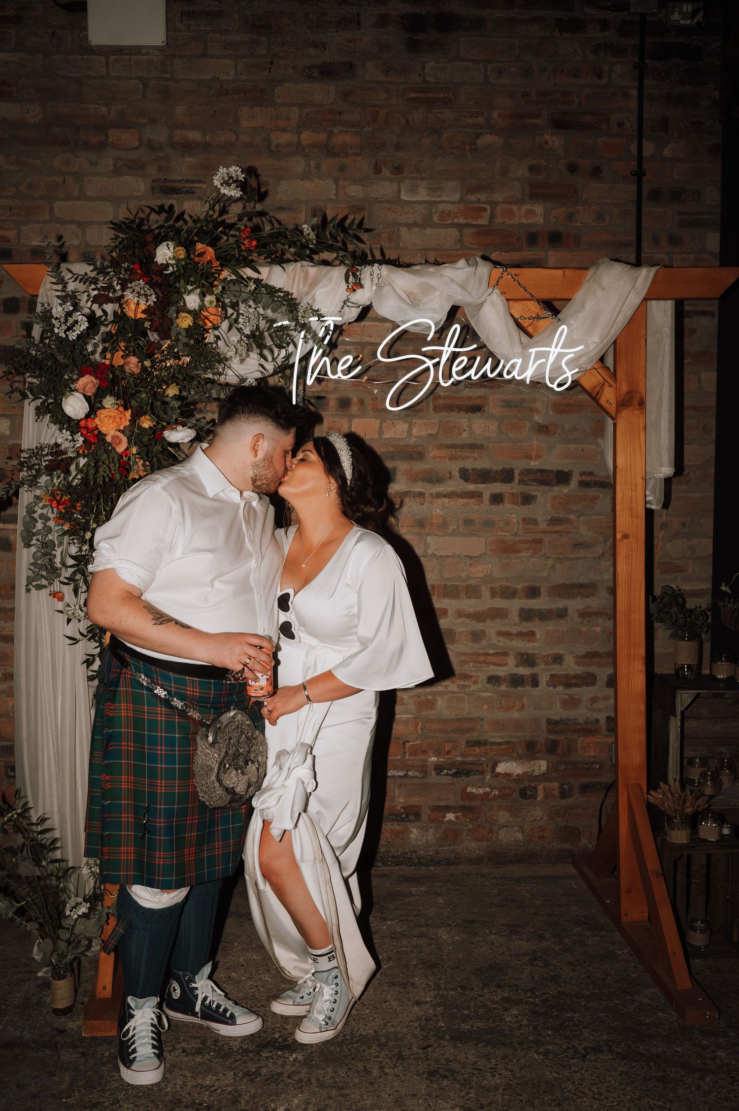 A couple poses under a floral arch with neon sign against exposed brick wall at Scottish-themed wedding.