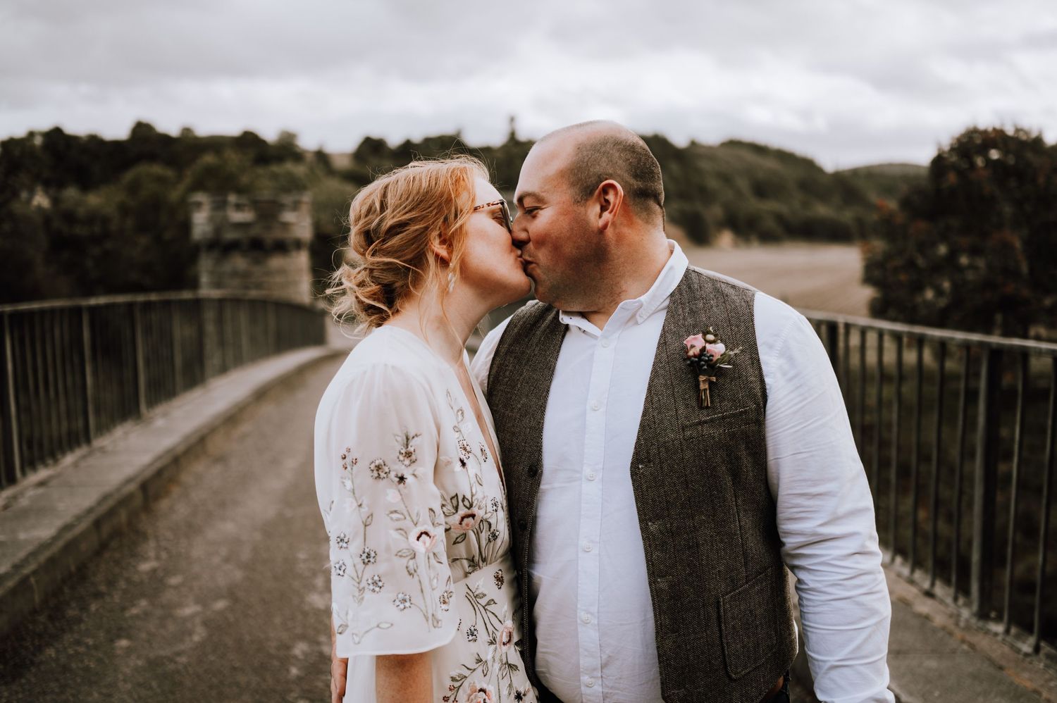 A tender kiss between newlyweds on a rustic bridge with rolling hills in the background.