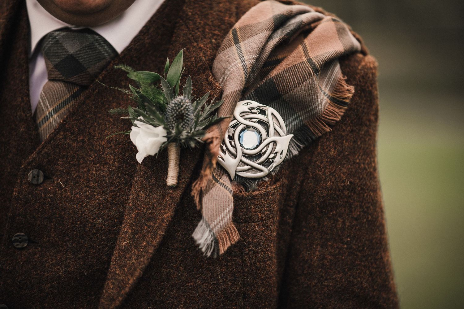 Close-up detail of white floral boutonniere and silver brooch on brown tweed wedding attire.