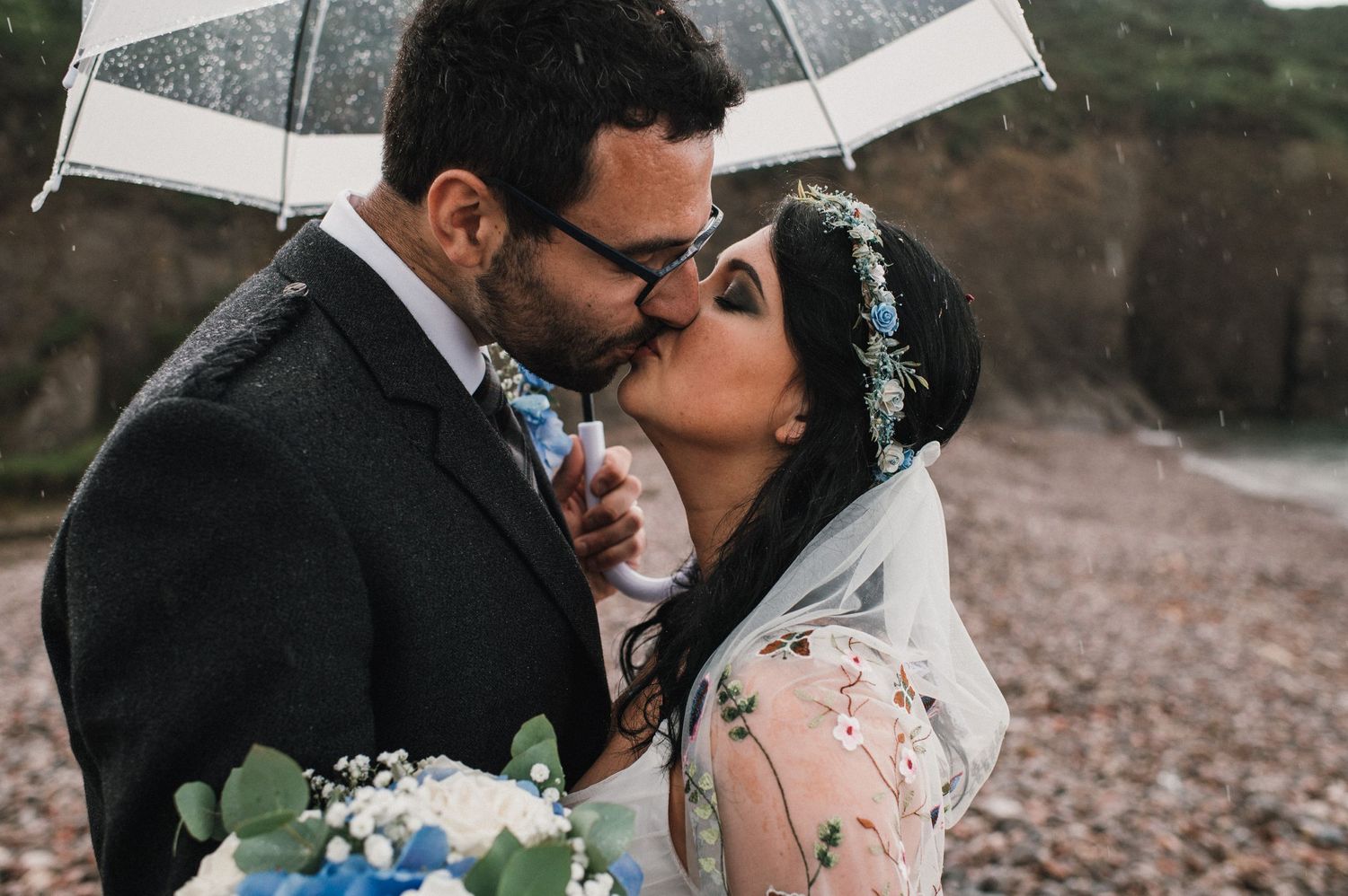A romantic moment between newlyweds sharing a kiss under an umbrella on a rocky beach.