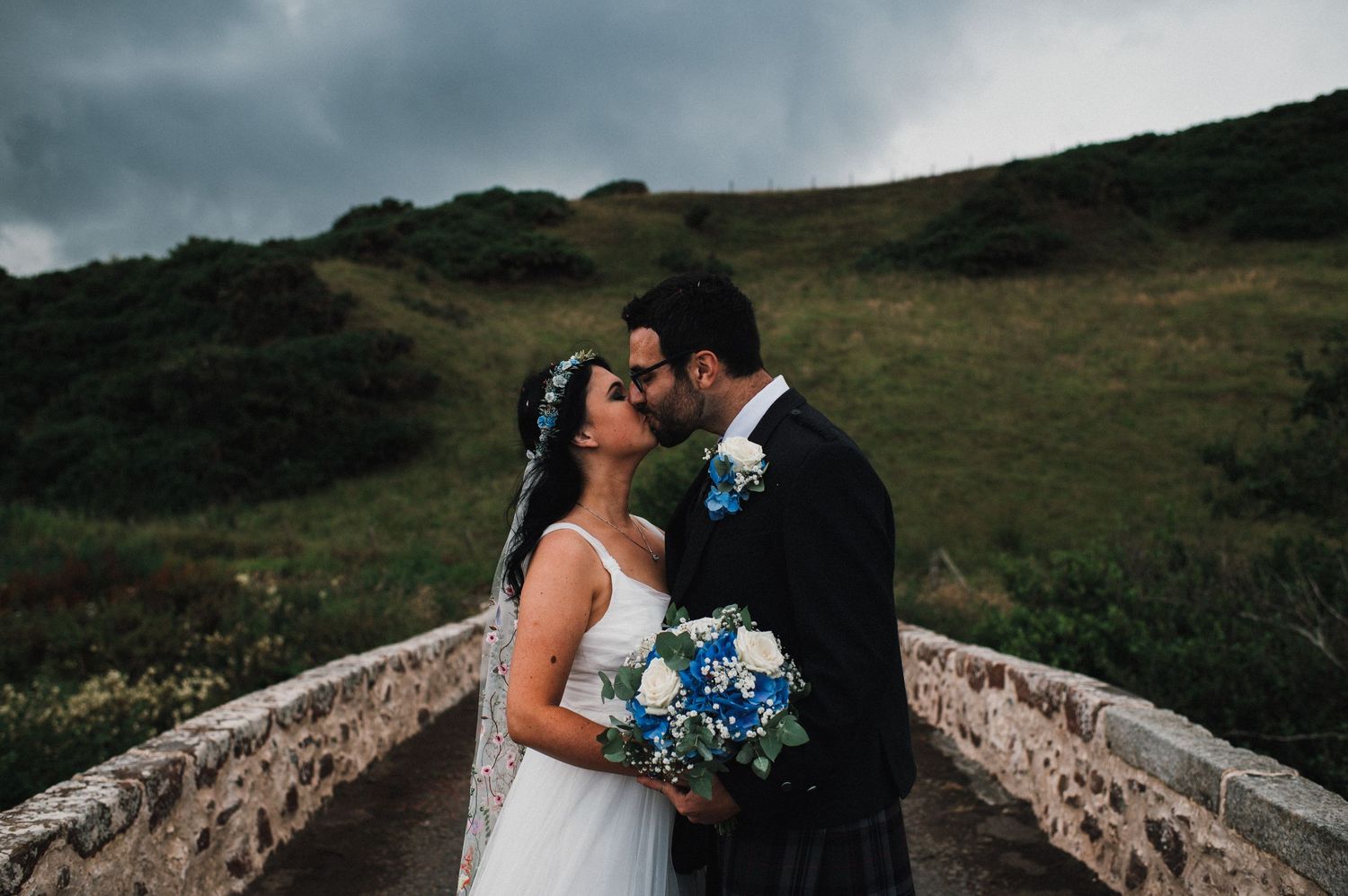 A romantic wedding moment captured on a stone bridge against moody hills and stormy skies.
