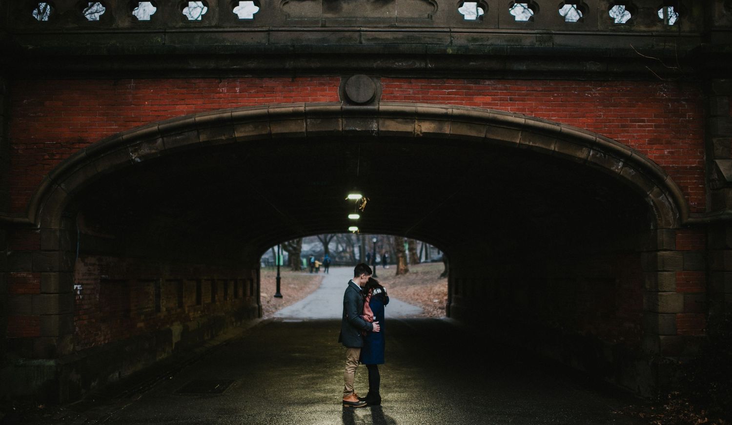 A couple embraces under a dark brick tunnel archway in an urban setting.