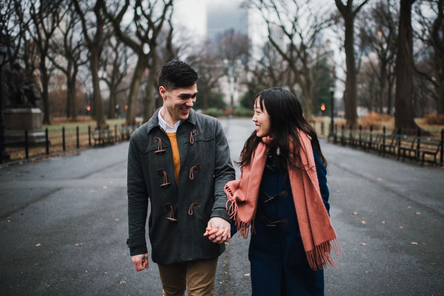 A couple walks hand in hand along a park path with bare winter trees and city buildings in the distance.