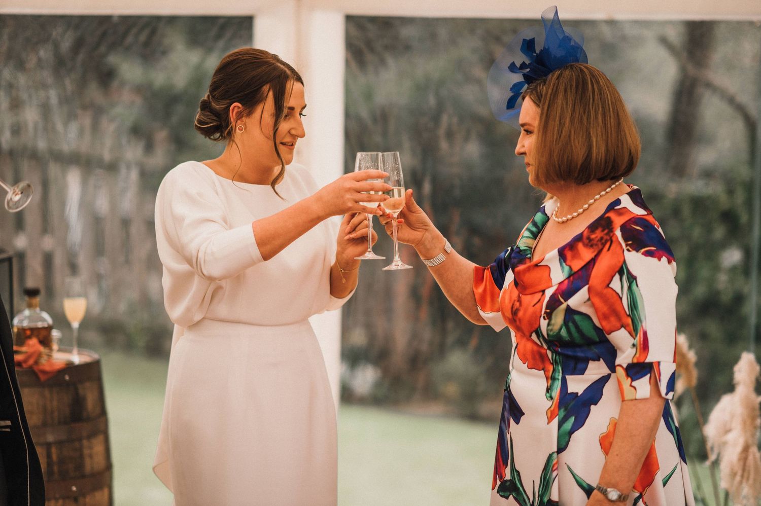 Two guests toast with champagne glasses at an outdoor event, one in white and one in colorful attire.