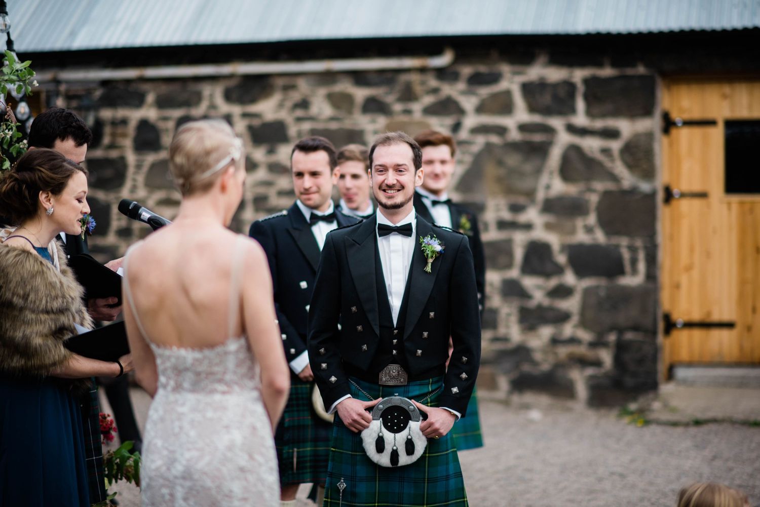 Guests in kilts and formal wear gathered around a stone building during a Scottish wedding.