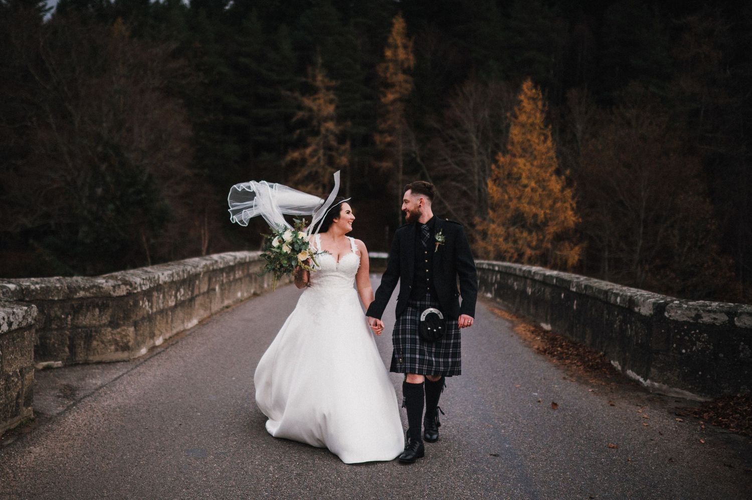 A couple walks along a stone bridge in autumn, with the bride in white and groom in traditional Scottish kilt.