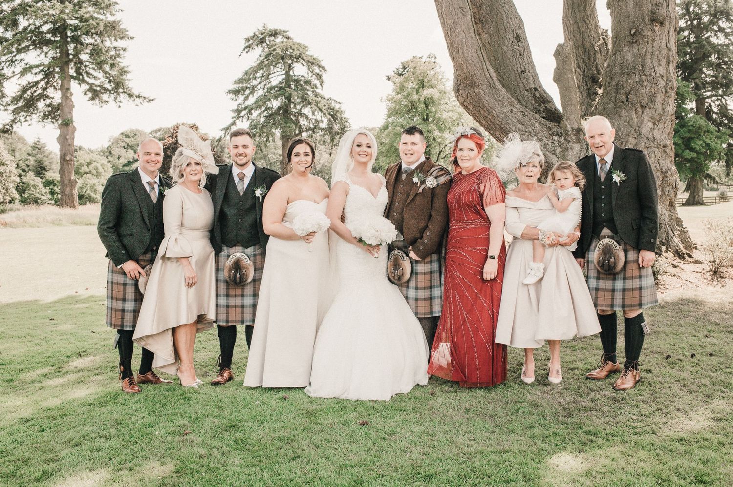 Wedding party in traditional Scottish attire poses outdoors on grass with mix of kilts and formal dresses.