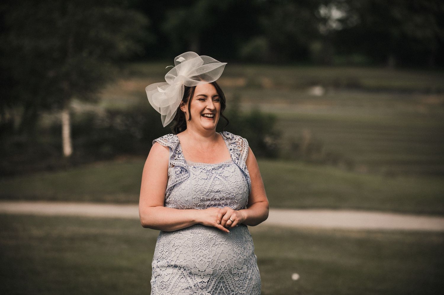 A person in a formal dress and fascinator smiling outdoors.