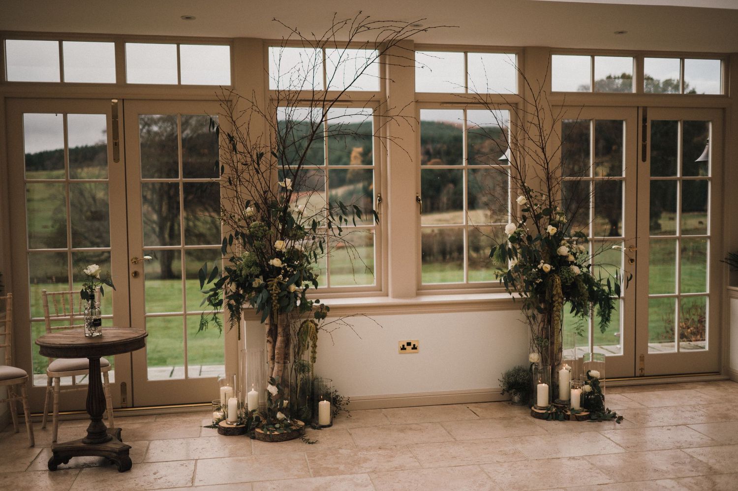 Large windows with floral arrangements and tall branches frame countryside view in elegant room.